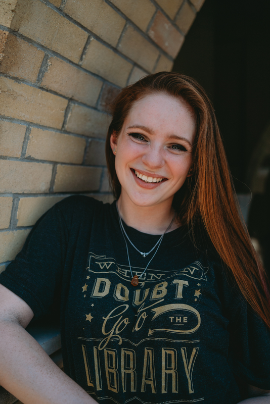 A headshot of Isabelle Antes with red brown hair, in a shirt that reads "When in doubt go to the library" in gold on a grey shirt. She leans against a tan brick archway.