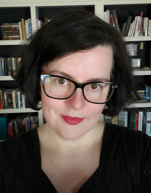 A headshot of Shelley Barba with dark hair and glasses in front of a bookcase